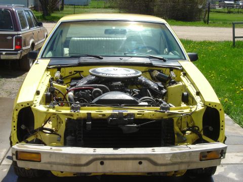 1979 Ford Mustang - Front view of a yellow 1979 Ford Mustang with its hood open, revealing the engine compartment. A brown station wagon is visible to the left.