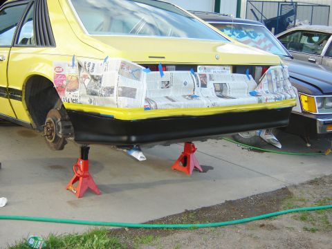 1979 Ford Mustang - A yellow car with racing decals is elevated on orange jack stands, showing its rear spoiler and suspension.