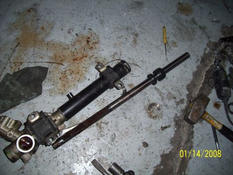 1995 Ford Probe - A disassembled steering rack and pinion assembly lies on a concrete floor, surrounded by tools and oil stains.