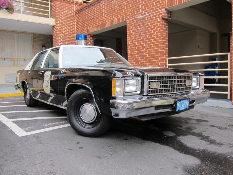 1991 Ford LTD Crown Victoria - black-and-white police sedan with a blue roof light bar parked at a curb in front of a red brick building.