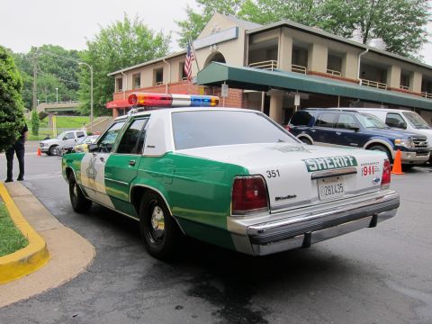 1991 Ford LTD Crown Victoria - Rear view of a green-and-white Ford Sheriff sedan with light bar parked outside a brick building flying an American flag.