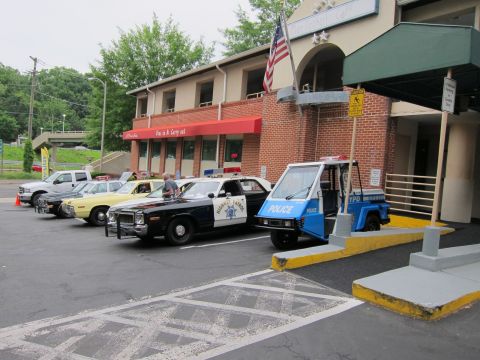 1991 Ford LTD Crown Victoria - line of vintage police vehicles, including a three-wheeled police parking cart, parked outside a brick building with an American flag.
