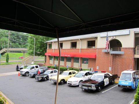 1991 Ford LTD Crown Victoria - row of parked vintage police cars and a three-wheeled police cart in a lot beside a brick building, viewed from under an awning.