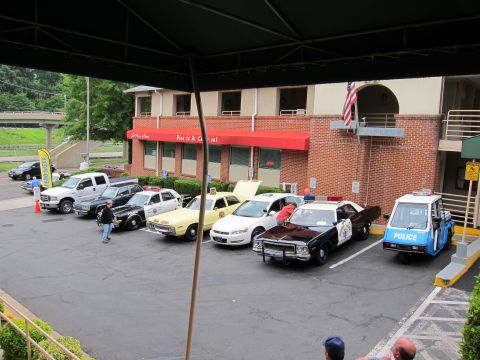 1991 Ford LTD Crown Victoria - row of classic police cars and a three-wheeled police cart parked in a lot beside a brick building, viewed past an umbrella pole.