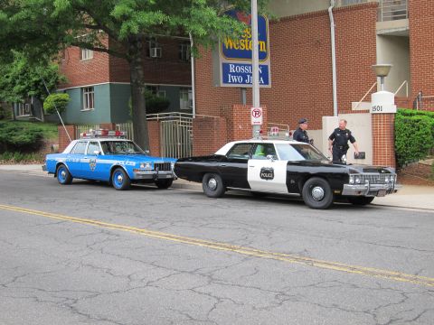 1991 Ford LTD Crown Victoria - blue police sedan and a black-and-white police car parked at a curb in front of a Best Western hotel as officers stand nearby.