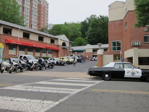 1991 Ford LTD Crown Victoria - black-and-white Highway Patrol sedan near a crosswalk with police motorcycles and vintage cars parked in a lot beyond.