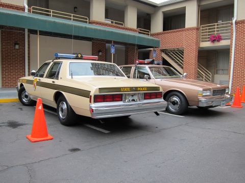 1991 Ford LTD Crown Victoria - tan State Police Caprice and a brown sedan parked side by side outside a brick motel with traffic cones nearby.
