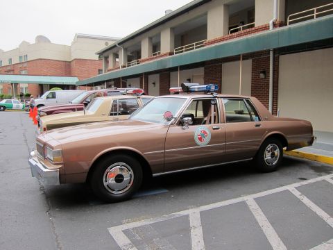 1991 Ford LTD Crown Victoria - brown Chevrolet Caprice police car with a roof light bar parked in marked spaces with other classic cars behind it.