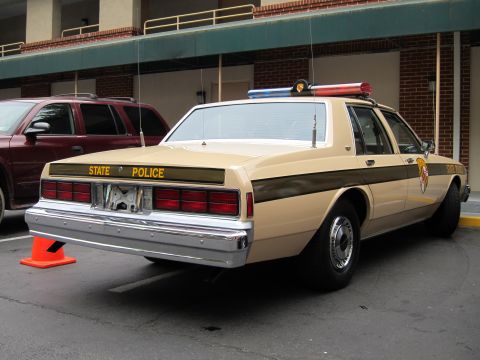 1991 Ford LTD Crown Victoria - Rear three-quarter view of a tan State Police sedan with a roof light bar, parked beside a maroon SUV outside a brick building.