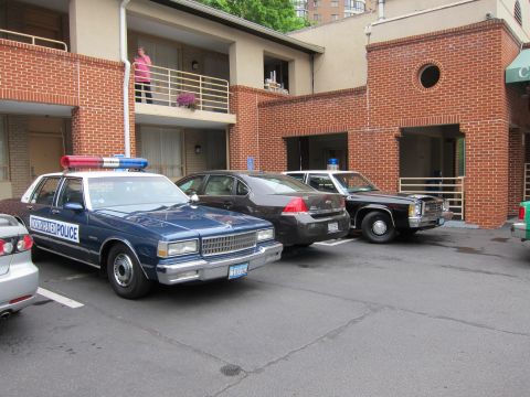 1991 Ford LTD Crown Victoria - dark blue police Caprice with roof lights and older sedans parked in marked spaces beside a brick motel building.
