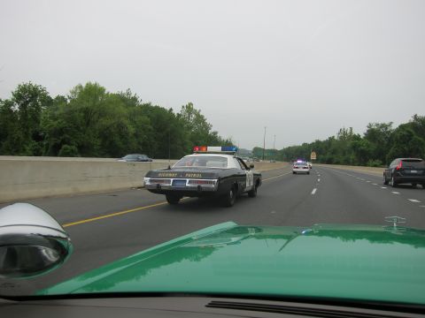 1991 Ford LTD Crown Victoria - black and white Highway Patrol cruiser drives down the highway ahead, viewed over the green hood of a following car.