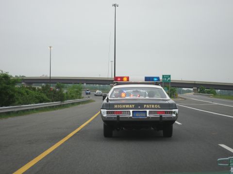1991 Ford LTD Crown Victoria - Rear view of a black Highway Patrol cruiser with roof lights driving toward an overpass on a multi-lane highway.