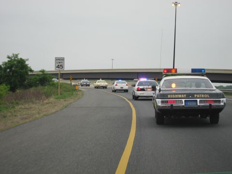1991 Ford LTD Crown Victoria - black Highway Patrol cruiser leads a procession of police cars along a curving road past a 45 mph speed limit sign.