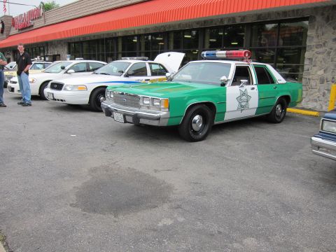 1991 Ford LTD Crown Victoria - green and white Ford police sedan with its hood raised parked in a line of cruisers outside a stone storefront.