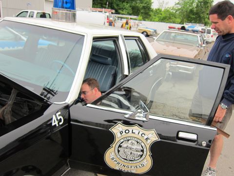 1991 Ford LTD Crown Victoria - Open driver door of a black Springfield Police sedan numbered 45, with a gold badge decal and a man seated inside.