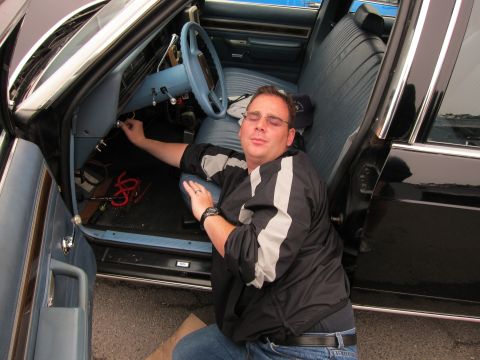 1991 Ford LTD Crown Victoria - man in a black and white striped jacket reclines on the ground reaching under the dashboard of a car with a blue interior.