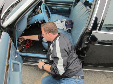 1991 Ford LTD Crown Victoria - man in a striped jacket kneels and reaches under the dashboard of a car with blue bench seats and exposed red wiring.