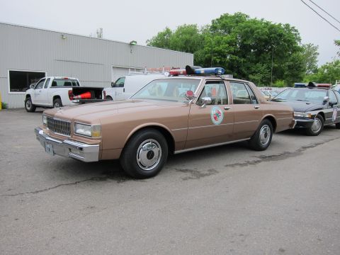 1991 Ford LTD Crown Victoria - tan Chevrolet Caprice police sedan with a roof light bar and door emblem parked in a lot beside a metal warehouse.
