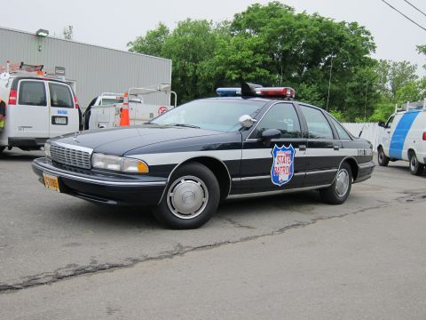 1991 Ford LTD Crown Victoria - black and silver Chevrolet Caprice State Patrol car with a roof light bar parked outside a warehouse near service vans.
