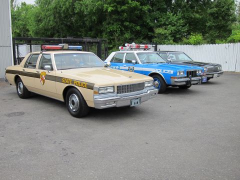 1991 Ford LTD Crown Victoria - lineup of three classic American police cars on pavement, a tan-and-brown State Police sedan in the foreground beside blue NYPD-style cruis…