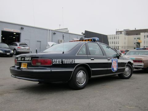 1991 Ford LTD Crown Victoria - Rear three-quarter view of a dark blue State Trooper sedan with roof light bar and shield decal, parked outside a repair shop.