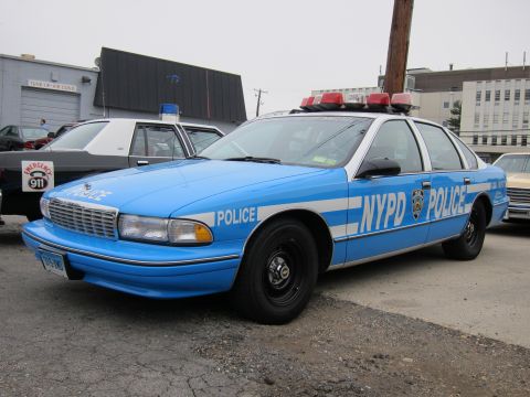 1991 Ford LTD Crown Victoria - light blue NYPD Police cruiser shown front three-quarter, with roof emergency lights, parked on pavement by buildings.