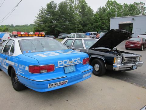 1991 Ford LTD Crown Victoria - Rear view of a blue NYPD Police cruiser with reward decal, parked beside a black sedan with its hood raised at a car show.