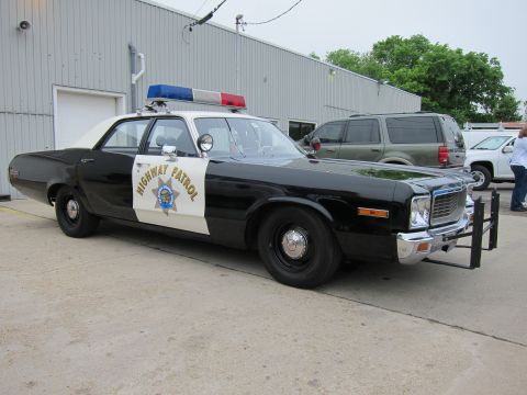 1991 Ford LTD Crown Victoria - black-and-white Highway Patrol sedan with spotlight and front push bar, parked on pavement beside a metal warehouse.