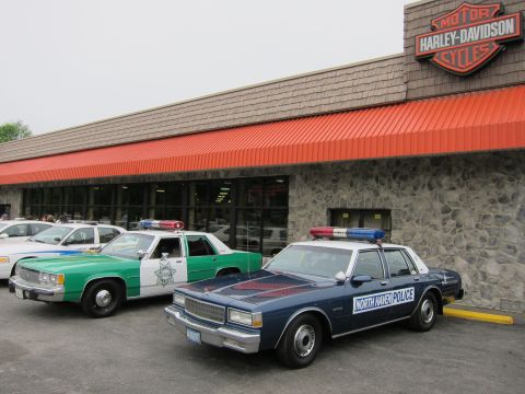 1991 Ford LTD Crown Victoria - green-and-white police sedan and a dark blue North Haven Police cruiser parked under an orange awning at a Harley-Davidson dealer.