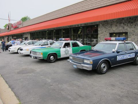 1991 Ford LTD Crown Victoria - long row of vintage police cruisers, including green-and-white and blue sedans, lined up outside a storefront with an orange awning.