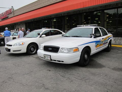 1991 Ford LTD Crown Victoria - white Ford police cruiser with blue striping parked beside a silver sedan outside a stone storefront with an orange awning.
