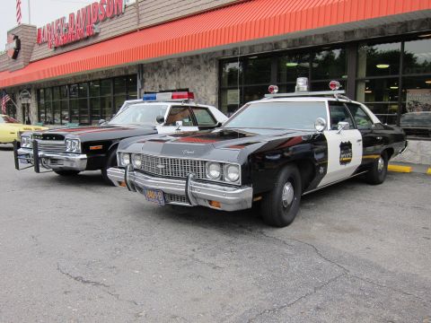 1991 Ford LTD Crown Victoria - black-and-white Chevrolet police sedan with chrome push bar parked beside a dark muscle-style sedan outside a storefront.