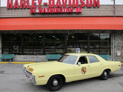 1991 Ford LTD Crown Victoria - yellow Police sedan with shield door decal parked in profile in front of the Harley-Davidson of Washington DC storefront.