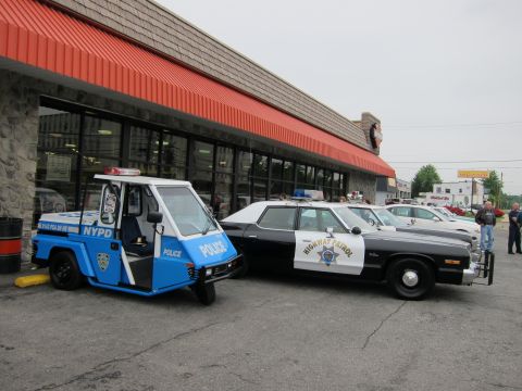 1991 Ford LTD Crown Victoria - small blue three-wheeled NYPD Police scooter parked nose-to-nose with a black-and-white Highway Patrol sedan outside a storefront.