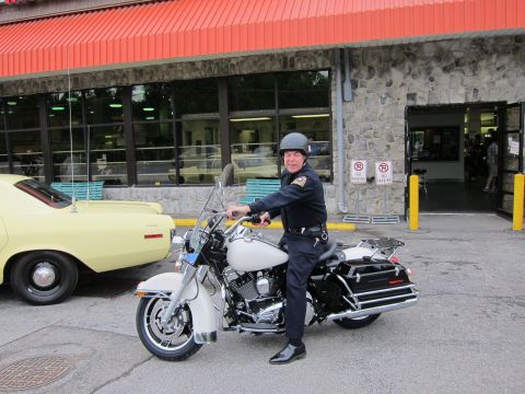 1991 Ford LTD Crown Victoria - uniformed police officer wearing a helmet sits astride a white Harley-Davidson police motorcycle on pavement outside a storefront.