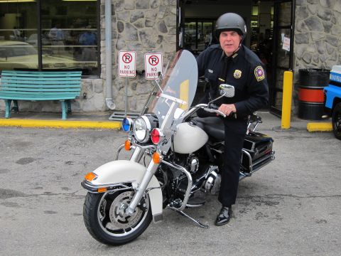 1991 Ford LTD Crown Victoria - uniformed officer in a helmet sits on a white police motorcycle with windshield, parked outside a stone-faced storefront.