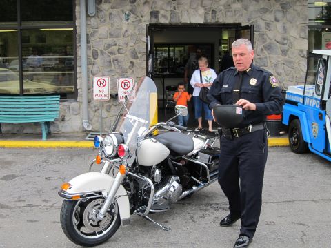 1991 Ford LTD Crown Victoria - uniformed police officer holds a clipboard while walking past a parked white police motorcycle outside a storefront entrance.