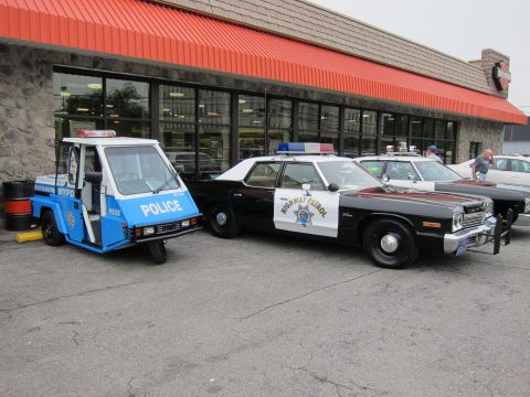 1991 Ford LTD Crown Victoria - blue three-wheeled NYPD Police scooter parked beside a black-and-white Highway Patrol sedan on pavement outside a storefront.