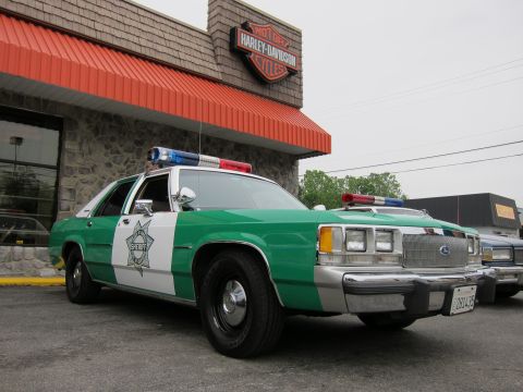 1991 Ford LTD Crown Victoria - Front three-quarter view of a green-and-white Ford Sheriff sedan with roof light bar parked outside a Harley-Davidson storefront.