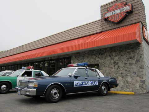 1991 Ford LTD Crown Victoria - dark blue North Haven Police sedan with roof light bar parked in front of a Harley-Davidson dealership with an orange awning.