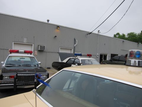 1991 Ford LTD Crown Victoria - Several police cars with red and blue roof light bars are parked tightly along a gray corrugated-metal building under an overcast sky.