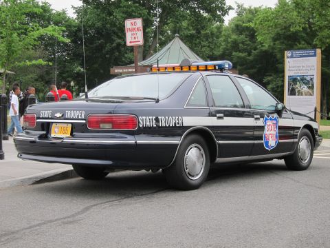 1991 Ford LTD Crown Victoria - dark State Trooper patrol sedan with a State Patrol shield and roof light bar is parked curbside as pedestrians pass.