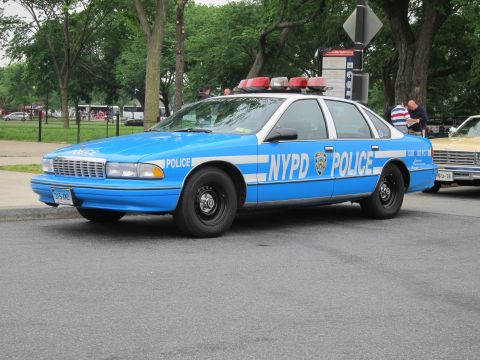 1991 Ford LTD Crown Victoria - blue-and-white NYPD Chevrolet Caprice police sedan with red roof beacons is parked on pavement in a park setting.