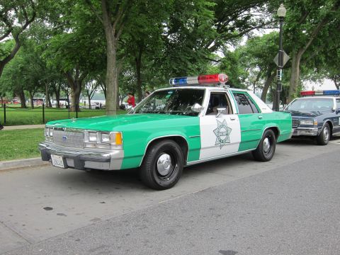 1991 Ford LTD Crown Victoria - green-and-white Ford Sheriff patrol sedan with red and blue roof beacons is parked among trees in a park.