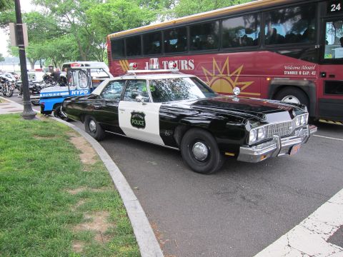 1991 Ford LTD Crown Victoria - black-and-white Chevrolet police sedan and a blue NYPD car are parked on a curving path beside a red sightseeing tour bus.