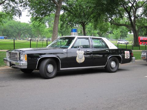 1991 Ford LTD Crown Victoria - black police sedan with a white roof, blue beacon and gold door shield is parked in profile along a park street.