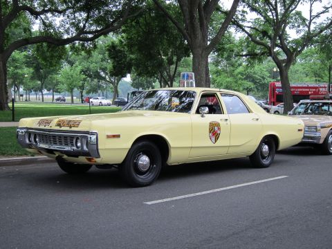 1991 Ford LTD Crown Victoria - pale-yellow State Police sedan with a round door shield is parked among trees in a park near a tour bus.