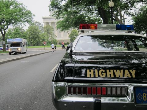 1991 Ford LTD Crown Victoria - Close-up of a black police cruiser's trunk with gold HIGHWAY lettering and roof light bar, parked along a tree-lined road near a stone monu…