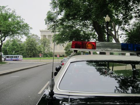 1991 Ford LTD Crown Victoria - View over the rear window of a black police cruiser showing a roof light bar and tall whip antenna, with a stone monument and bus in the ba…