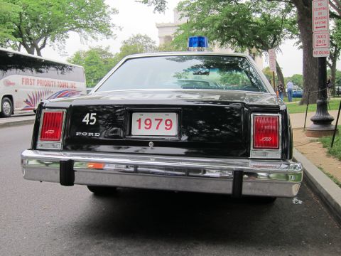 1991 Ford LTD Crown Victoria - Rear view of a black Ford LTD police sedan marked 45 with a 1979 license plate and a single blue roof beacon, parked curbside near a tour b…